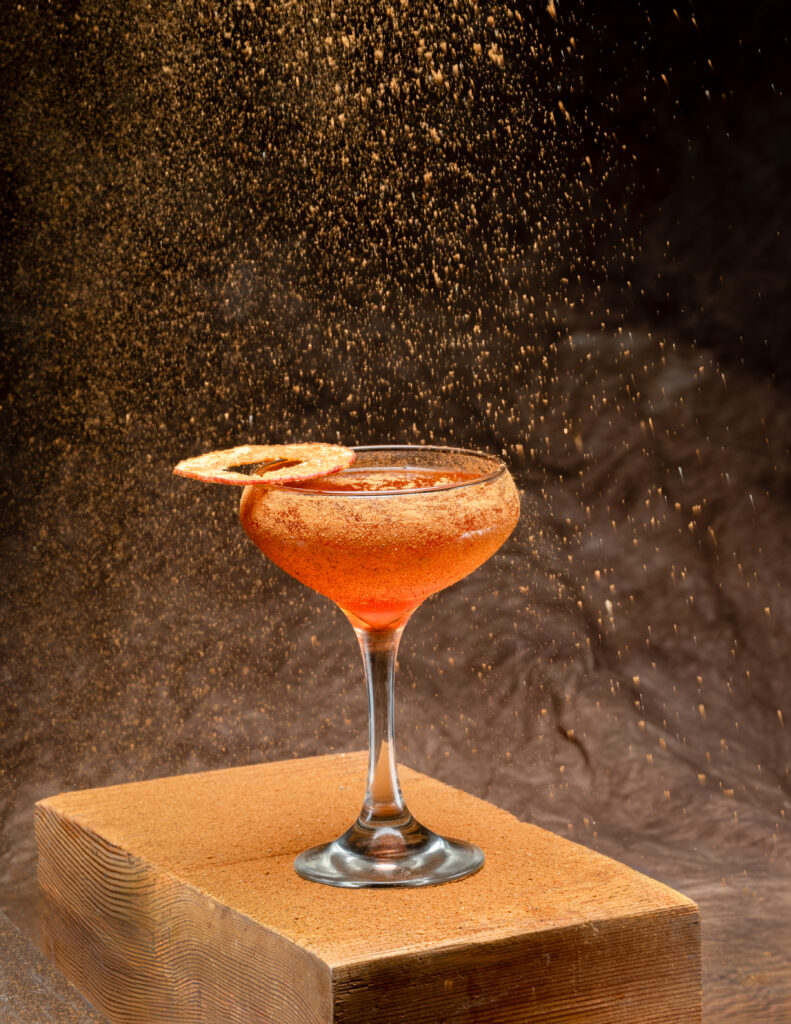 A cocktail in an orangish hue sits in a cocktail glass topped with an apple chip and dusted in spice for our Sand of Arrakis Dune cocktail.