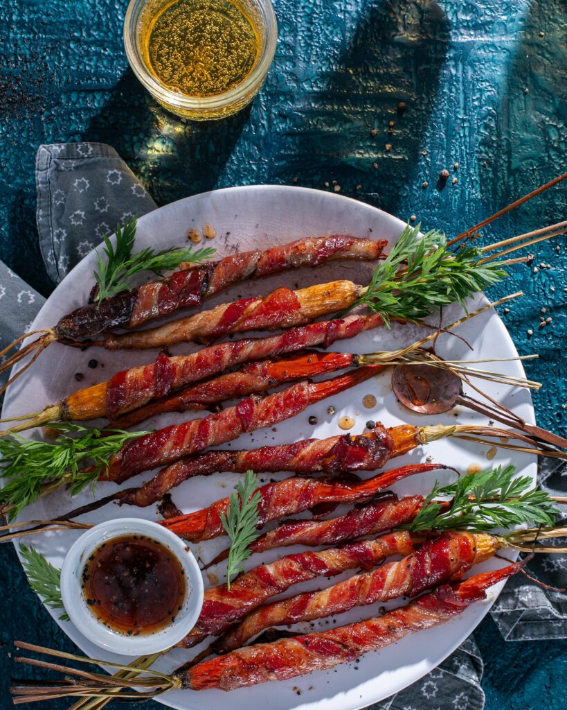 On a white plate sits a bundle of maple-glazed bacon-wrapped carrots with a maple dipping sauce to the side and a glass of beer above the plate.