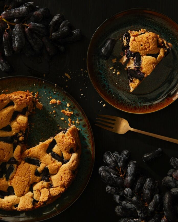 A black grape torte sits on a plate on a table while a slice sits nearby on another plate and a fork is in between the two.