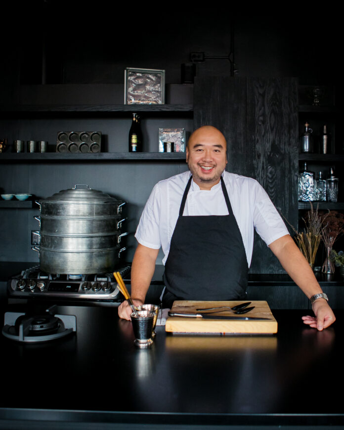 Chef Roger Li stands in front of a black decorated kitchen in a black apron with a cutting board in front of him.