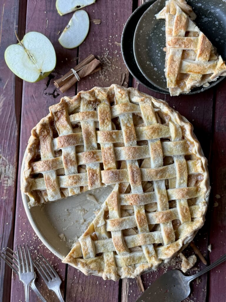 A traditional apple pie with a lattice top crust on a worn red wood surface with a sliced apple, cinnamon sticks, a slice of pie and silver forks.