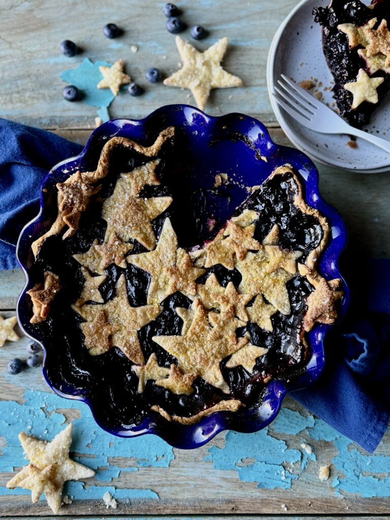 A cherry blueberry pie with stars as a top crust, in a cobalt blue, scalloped edge pie dish sitting on a blue cotton towel on a rustic wooden surface with chipped light blue paint