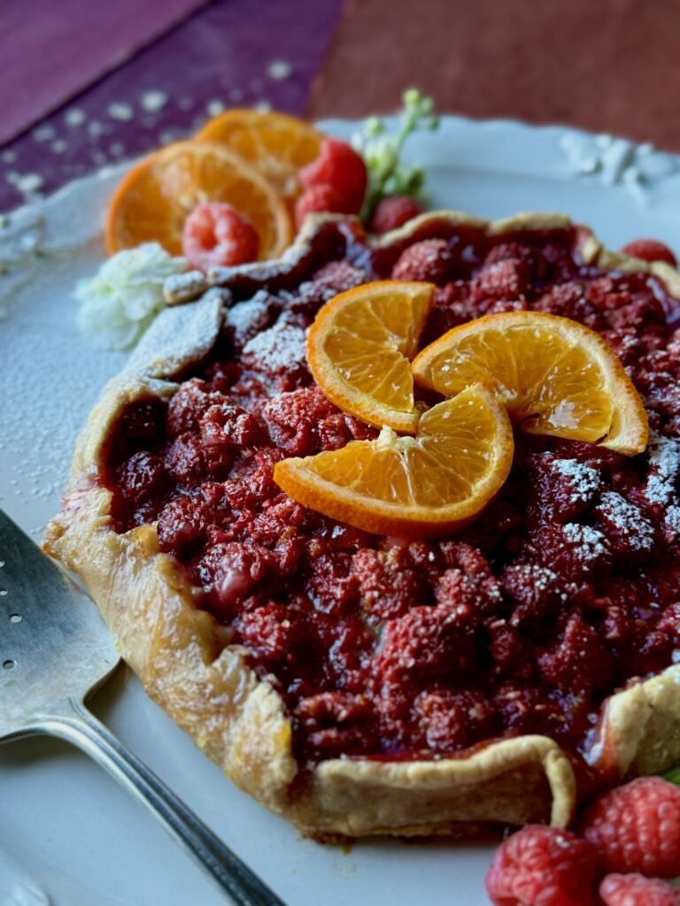 a lookin at a raspberry orange galette with sliced oranges, white flowers, and raspberry garnish on a white plate.