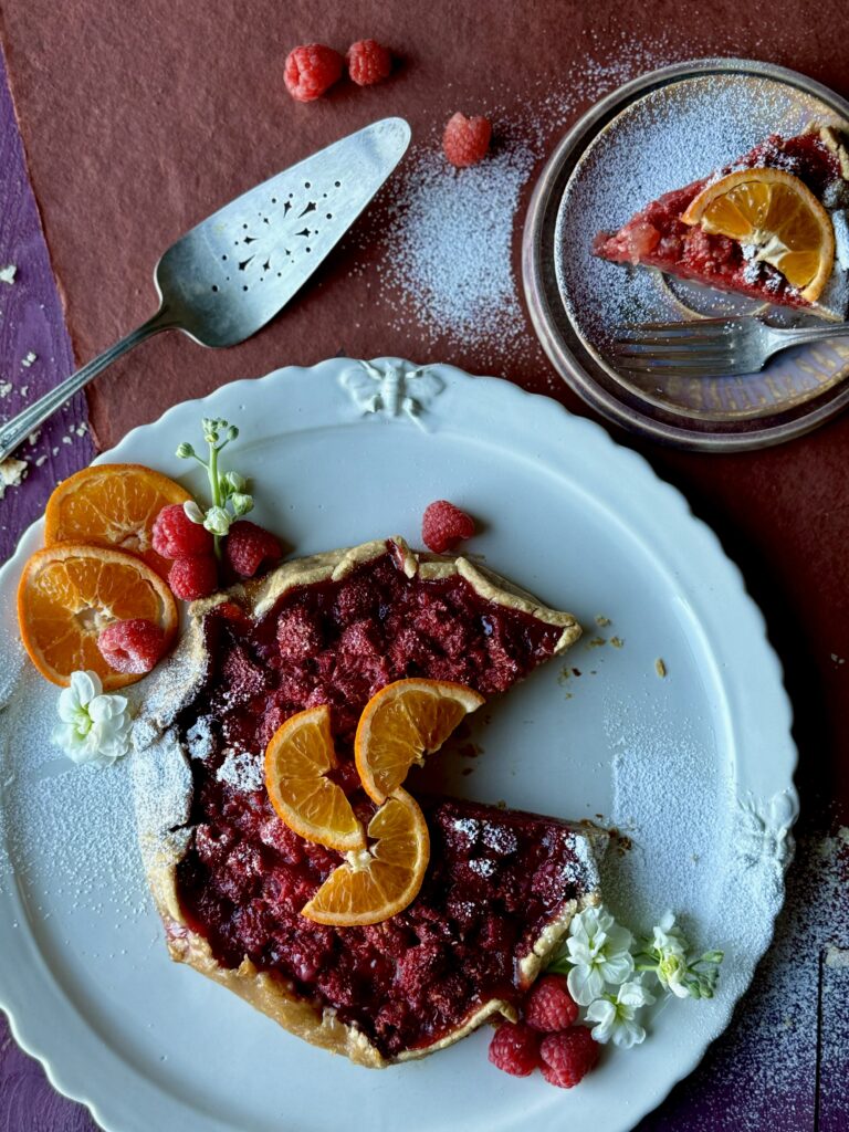 A rustic Raspberry Orange Galette on a round white plate with sliced oranges and white flowers, a pie server and a slice of the galette on a plate in the upper right corner.