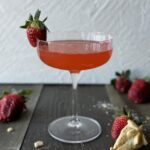A strawberry rhubarb pie cocktail in a clear glass with pie crust strawberries and fresh strawberry garnish on a wooden surface with a white textured background