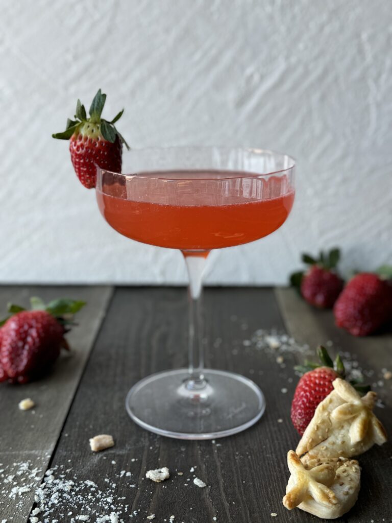 A strawberry rhubarb pie cocktail in a clear glass with pie crust strawberries and fresh strawberry garnish on a wooden surface with a white textured background