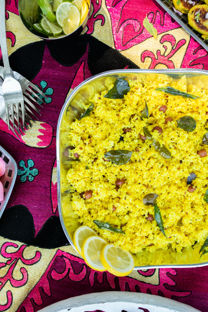 lemon rice with toasted peanuts served in a steel tray with three lemon slices in the corner. The tray placed on a colorful cloth with 4 steel forks