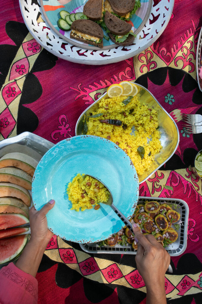 A person holding a blue plate scoops Lemon Rice with Toasted Peanuts onto it, above an array of picnic dishes all on a pink blanket.
