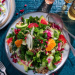 A plate with a salad on it has plenty of kale and citrus fruits like orange slices with drinks in the top right corner of the photo and more plates and forks to the left.