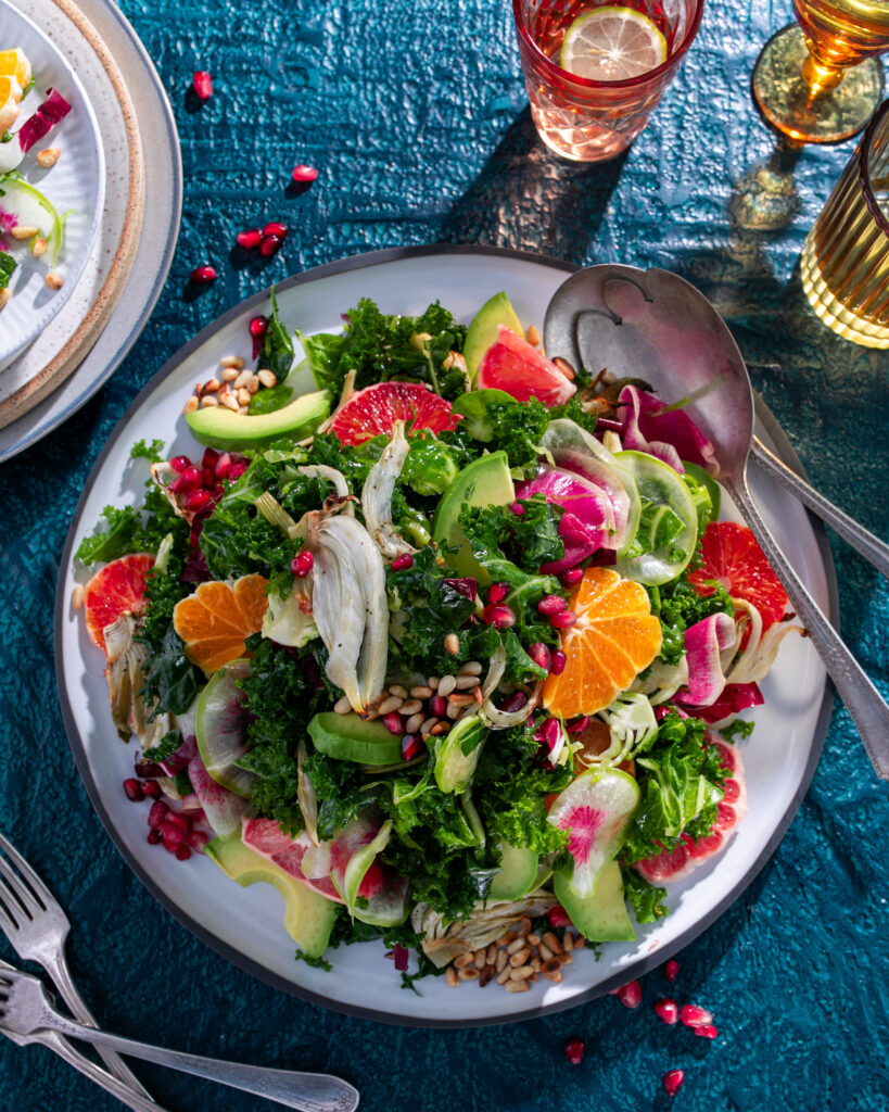 A plate with a salad on it has plenty of kale and citrus fruits like orange slices with drinks in the top right corner of the photo and more plates and forks to the left.