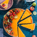 A big orange round tart littered with tomatoes and lime slices surrounded by individual tomatoes and limes and a slice of the tart on a pink plate.