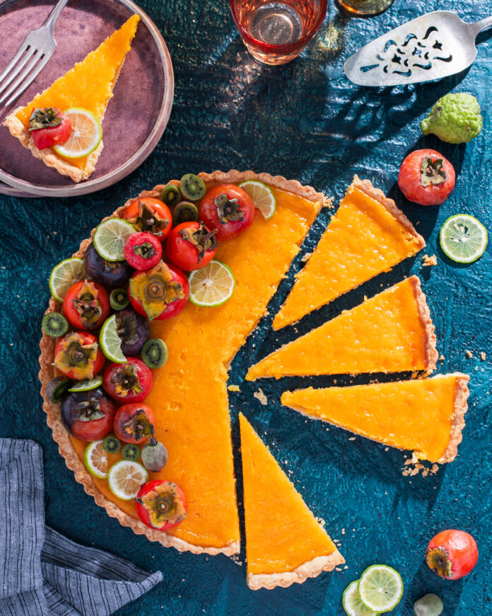 A big orange round tart littered with tomatoes and lime slices surrounded by individual tomatoes and limes and a slice of the tart on a pink plate.