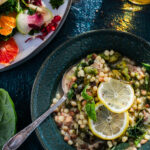 A bowl of soup with little round noodle balls, greens, chicken, and lemon slices on top sits in a dark teal bowl with a winter citrus salad sitting nearby.