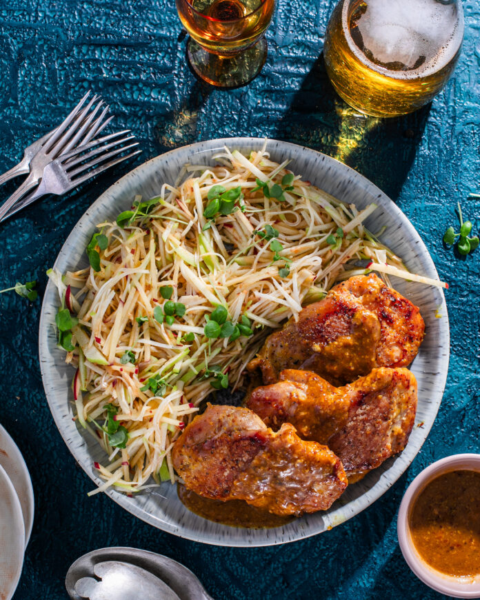 A plate holds a dinner of honey mustard chicken and a celery root slaw with two drinks sitting above the plate and two forks to the left.
