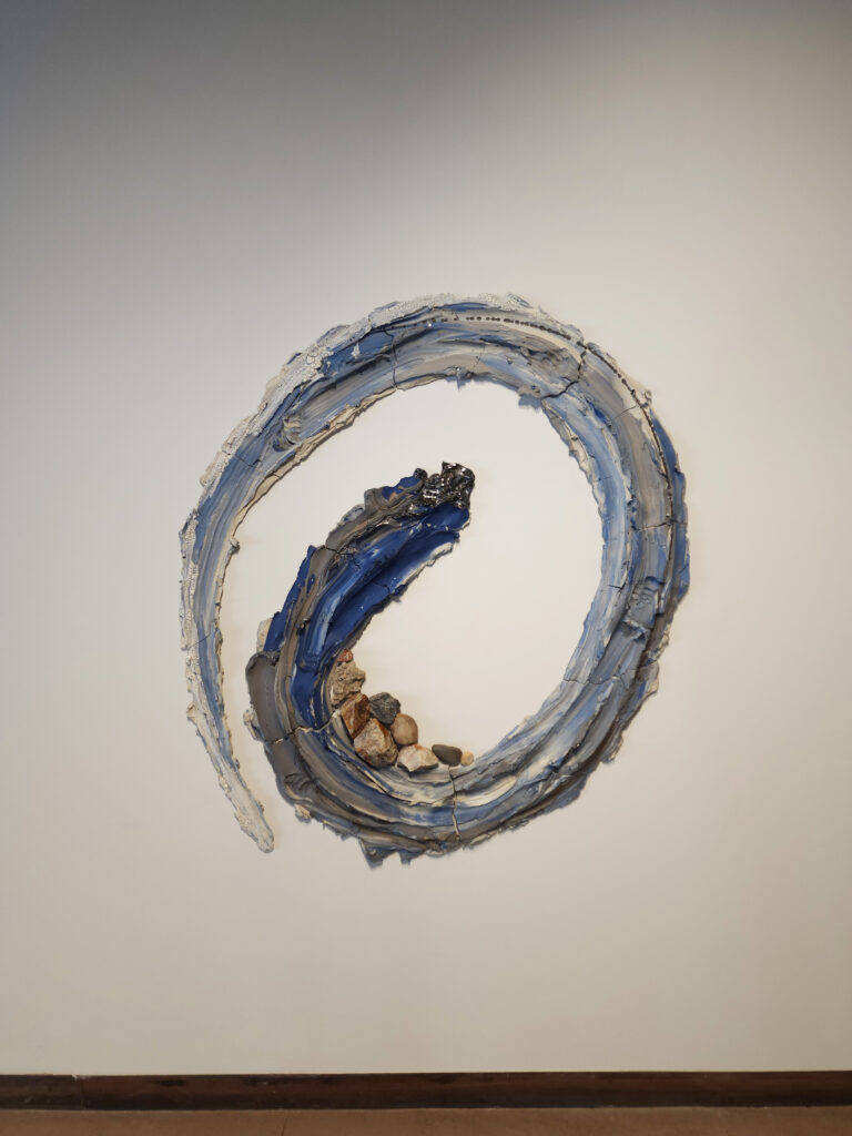 The work of Brie Ruais' Oneness, a blue and brown swirled clay is smeared in a spiral on the wall of Contemporary Craft with loose stones sitting in the middle.