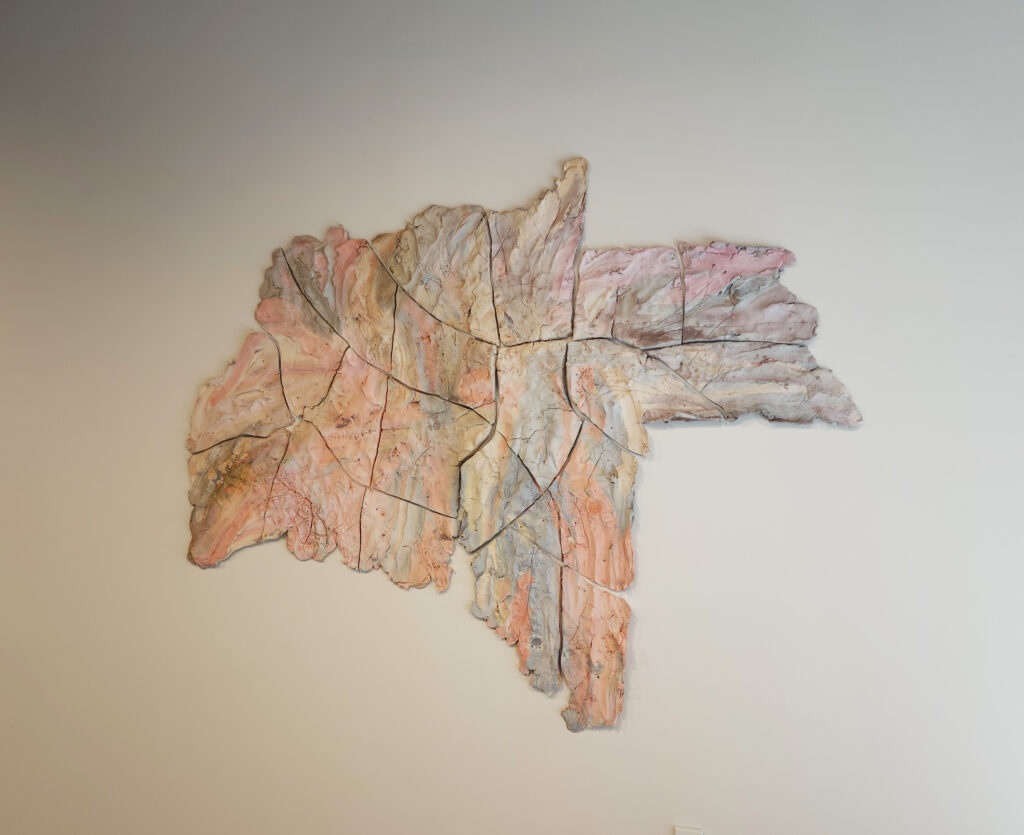 Brie Ruais Oneness displays a piece of clay smeared on the wall in a messy shape colored with light orange, purple, blue, and grey.