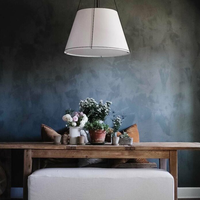 An interior shot of a dark washed design trend wall with a wood table and grey ottoman in front of it and white light over the table.