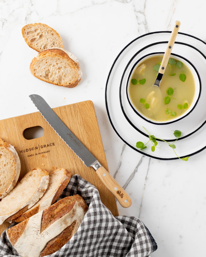 A wood cutting board holds slices of bread and a knife as a soup bowl sits nearby.