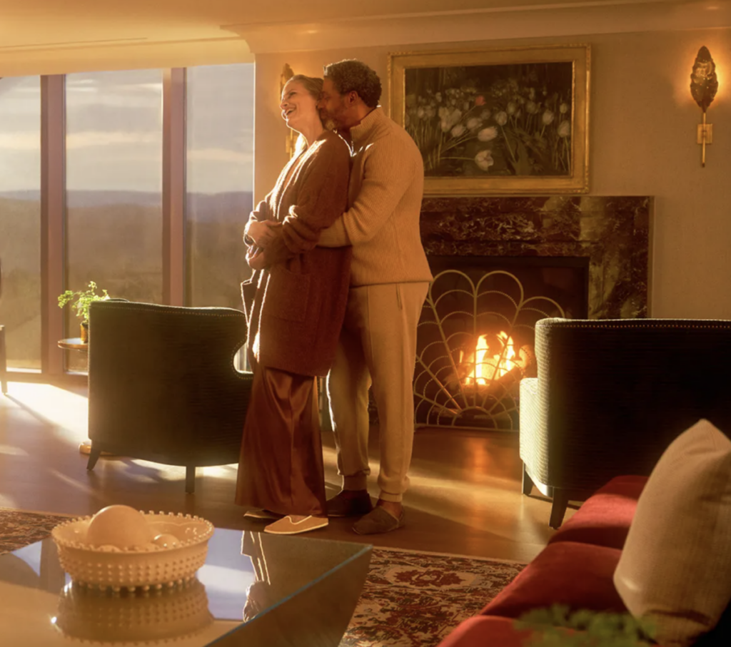 A couple stands in a Valentine's Day Hotel package suite at Nemacolin with a fireplace in the distance as they look out the long windows.
