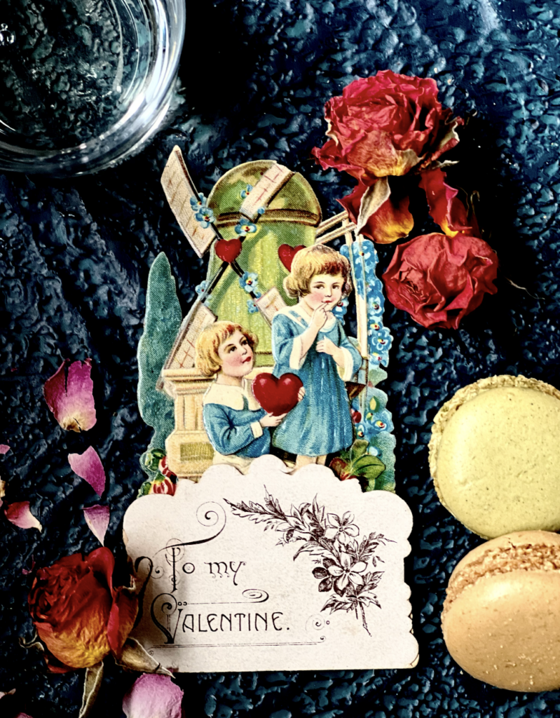 A vintage valentine with two little children in teal dresses sits on a blue background surrounded by dried flowers and macarons. 