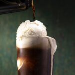 A closeup look in photo of a Guinness Beer Float with vanilla ice cream and a can of beer being poured over the top of a ball of creamy white ice cream.