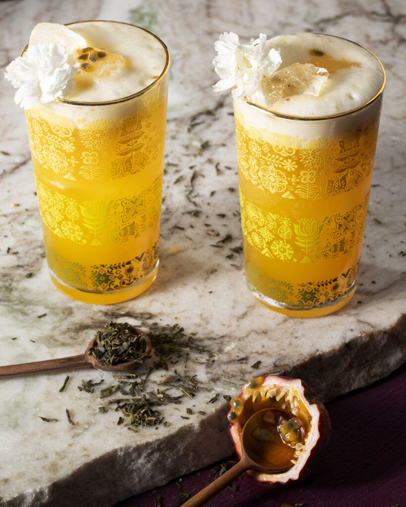 Two glasses filled with a Francis Forever Valentine's Day Mocktail with a spoon of herbs nearby and a piece of passionfruit in front of the glasses.