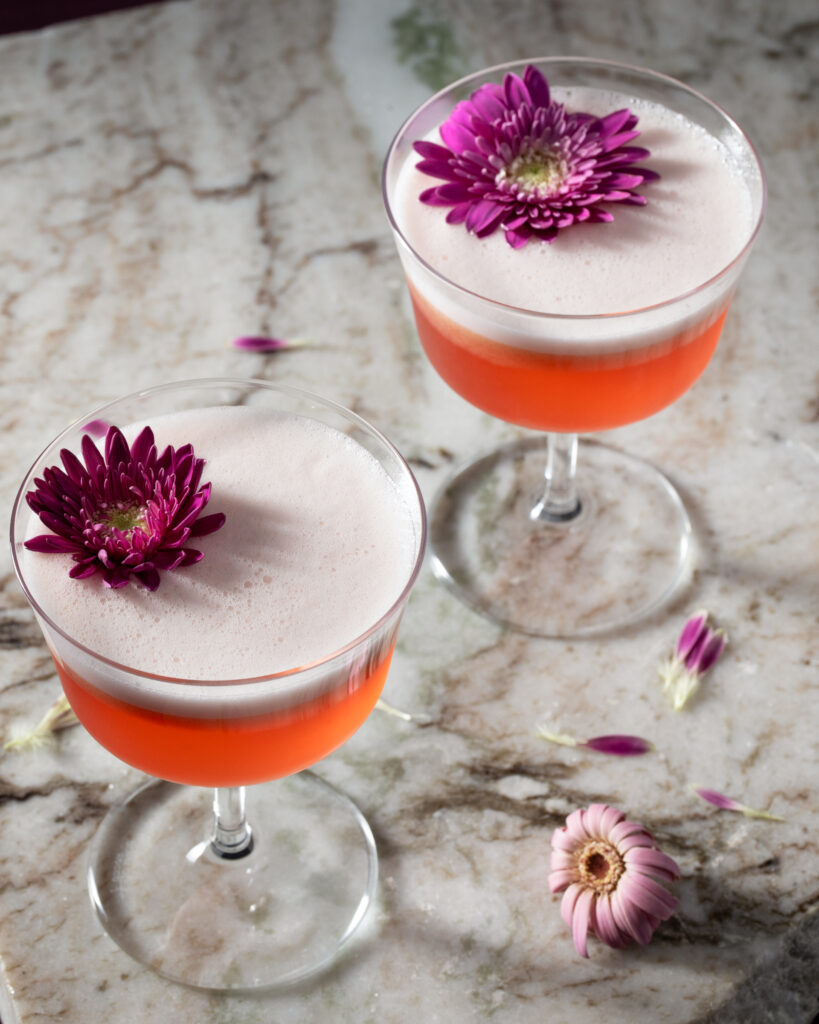 Two coupe glasses hold slightly red The Archer Valentine's Day Cocktail with an edible purple flower on top.