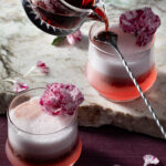 Two small cocktail classes hold a valentine's day cocktail in a red color with a foam on top and edible flowers as a garnish.