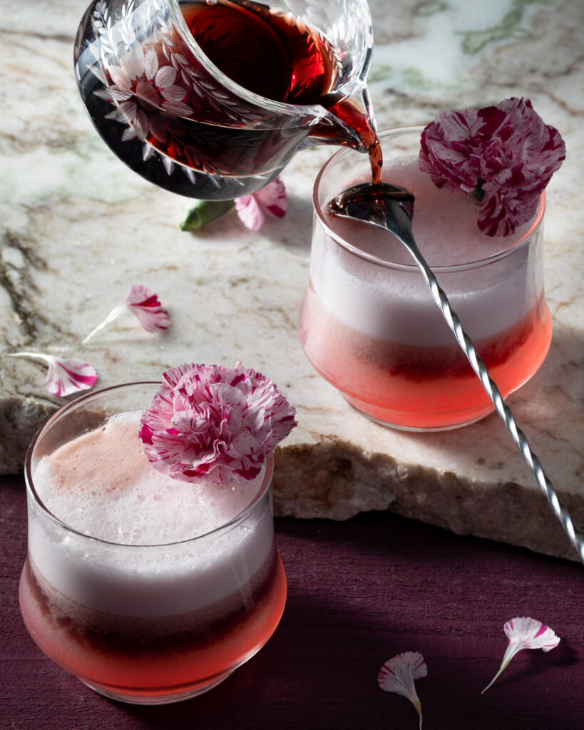 Two small cocktail classes hold a valentine's day cocktail in a red color with a foam on top and edible flowers as a garnish.