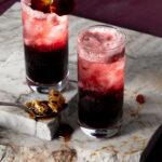 Two glasses sit on slabs of marble with deep red liquid at the bottom and topped with a red edible flower to represent the Chateau Lobby #4 Valentine's Day mocktail.