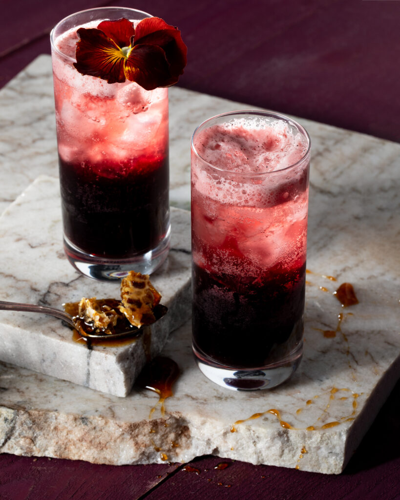 Two glasses sit on slabs of marble with deep red liquid at the bottom and topped with a red edible flower to represent the Chateau Lobby #4 Valentine's Day mocktail.