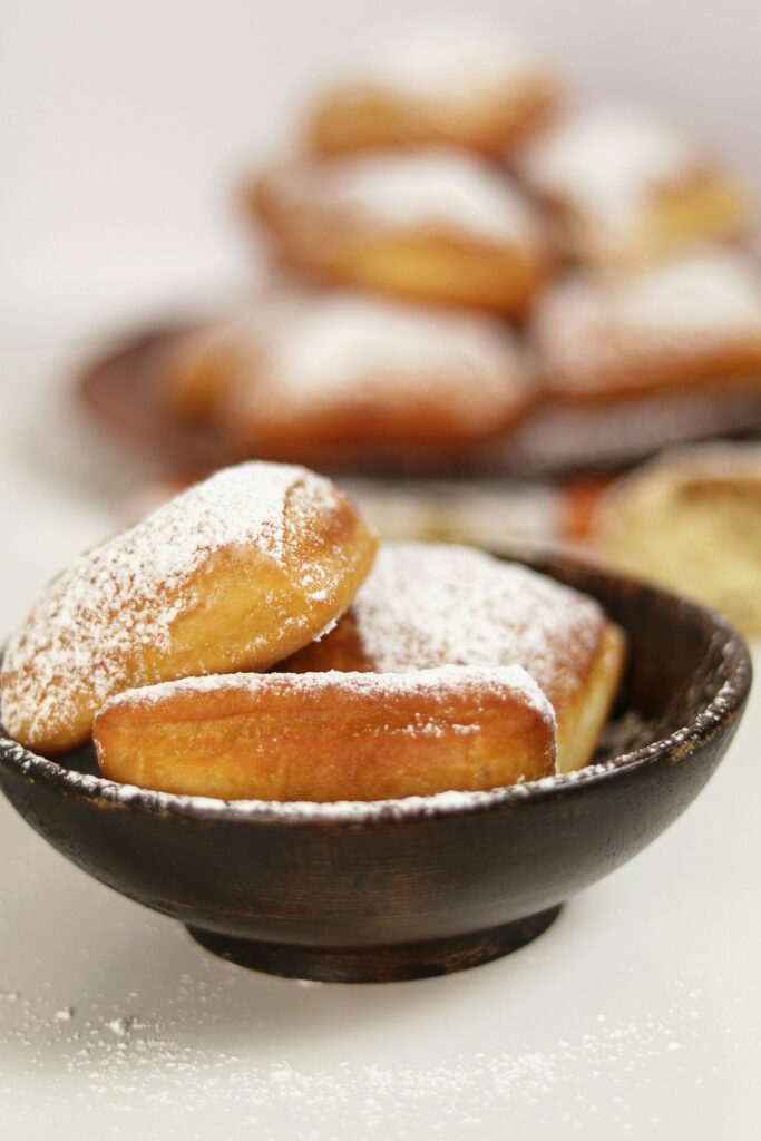 Powdered sugar topped beignets sit in a black bowl while a plate of beignets sits unfocused in the back.