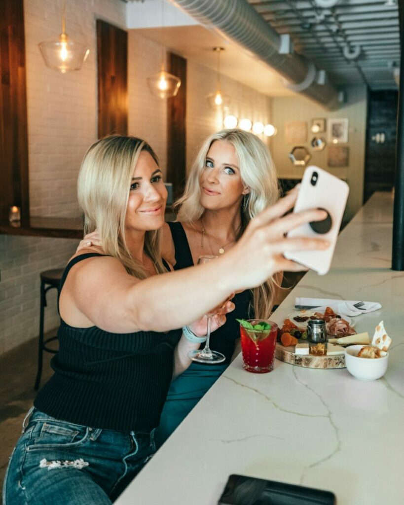 two women stop during brunch for a selfie at the brunch counter as their drinks and food sit in front of them.
