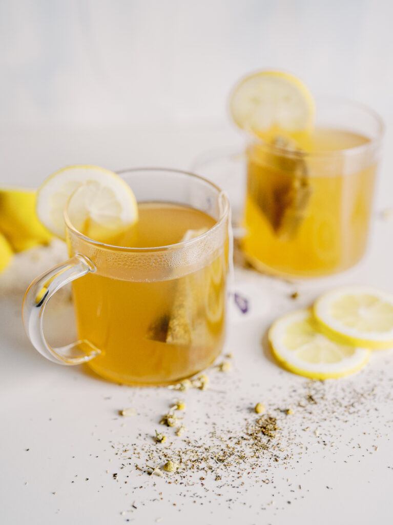 Two mugs filled with a golden hot honey toddy liquid with a tea bag in each and lemon slice on the rim. They're on a white background surrounded by spices and lemon slices.