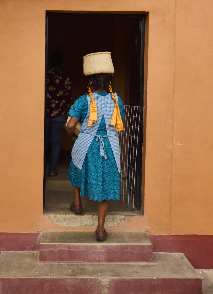 A person dressed in all blue steps out of a house with a basket on their head.