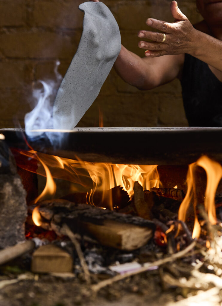 A persons hands flip a sheet of maza over top of a fire under a cooking stone.