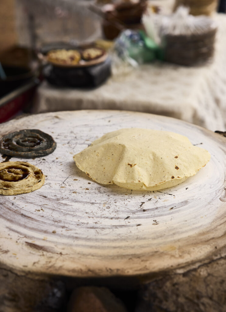 A stone holds a puffed up tortilla made of masa with a table in the background.