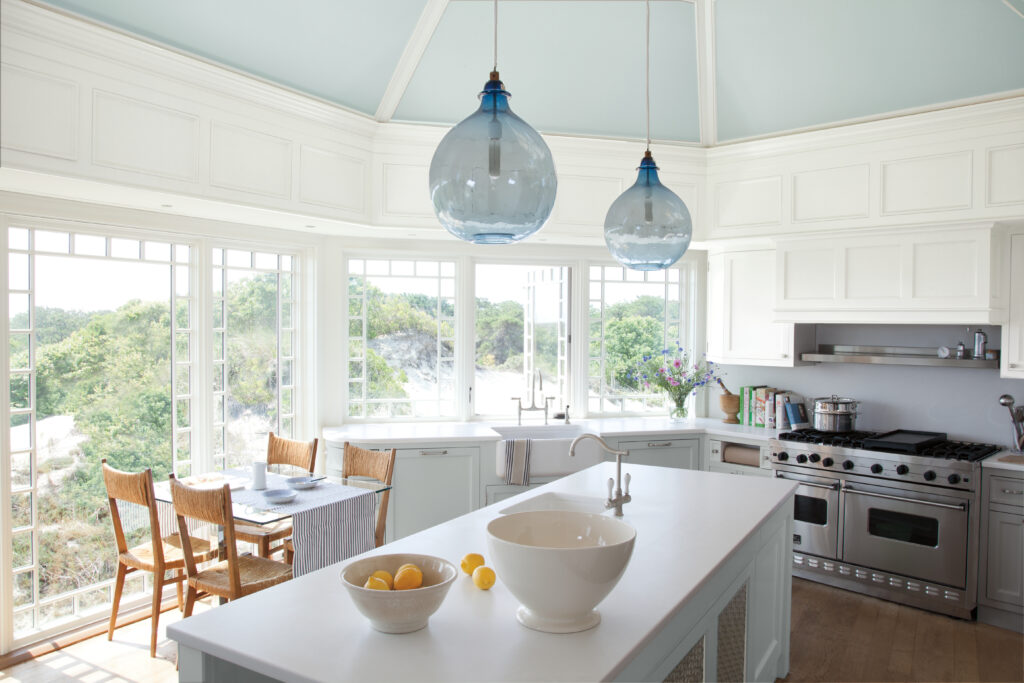 An open kitchen with plenty of windows and white painted appliances with two blue glass lights hanging above the counter.