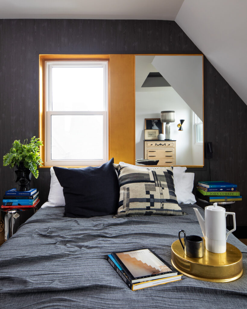 An interior shot of a bedroom with dark navy sheets, dark walls, and a splash of gold around the window.