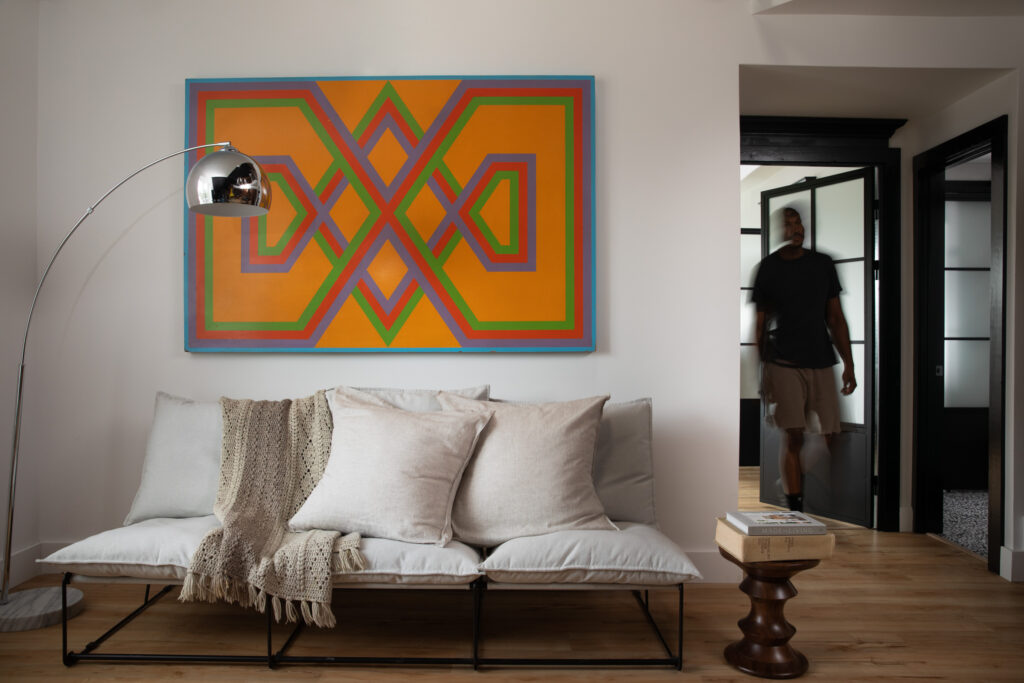 A living room holds a minimalist white couch with pillows and a blanket as an orange painting sits above it. To the left Cinque Cerra-Saunders walks through a doorway, blurred.