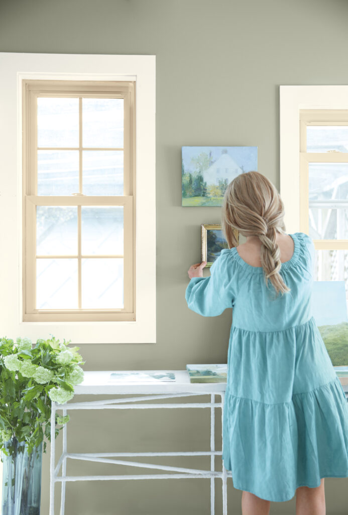 A woman in a blue dress hangs a painting against a sage colored wall with windows outlined in beige.