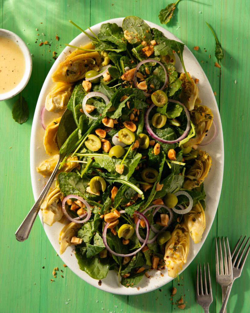 Baby Kale Salad with Citrus-Marinated Artichoke Hearts and Lemon Vinaigrette on a white plate with a fork in the salad and a container of dressing to the left.