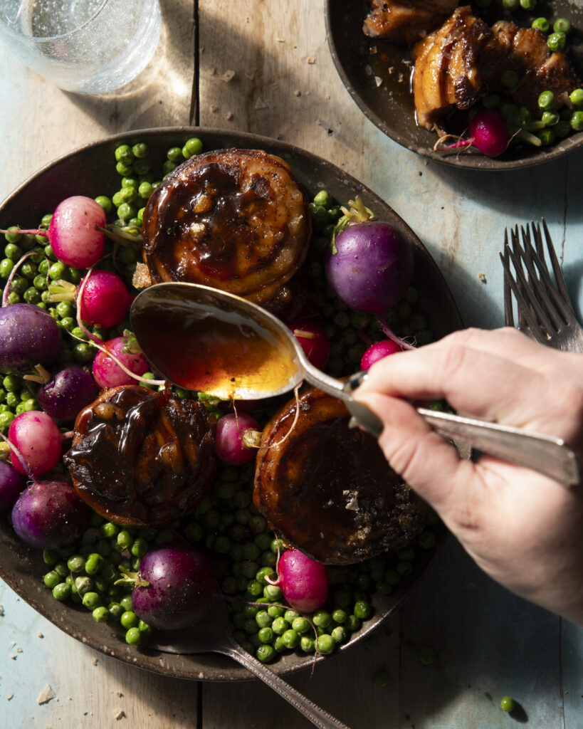 A silver spoon with pours raw apple cider over a bowl of braised pork belly and fresh vegetables.
