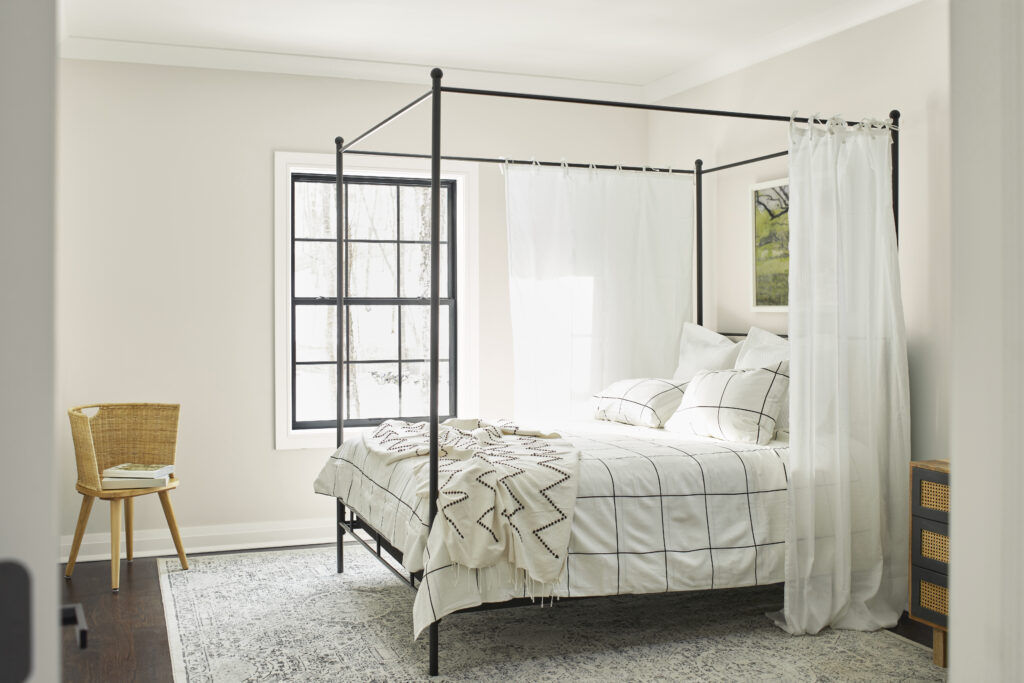 A four poster bed set sits in a white/beige spring paint colored room with a window letting sunlight shine through.