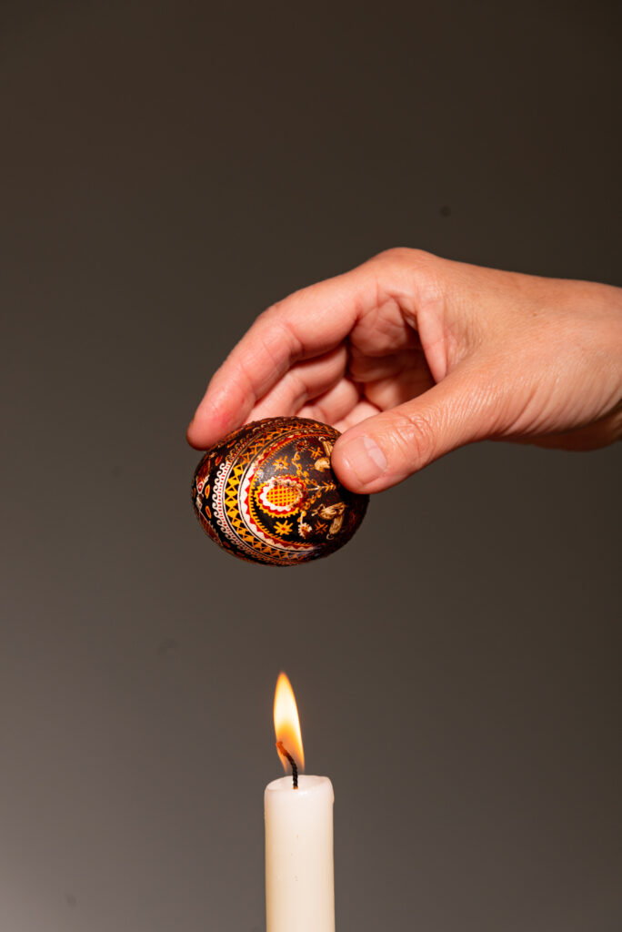 A woman uses a candle flame to melt the wax off of a Pysanky egg.