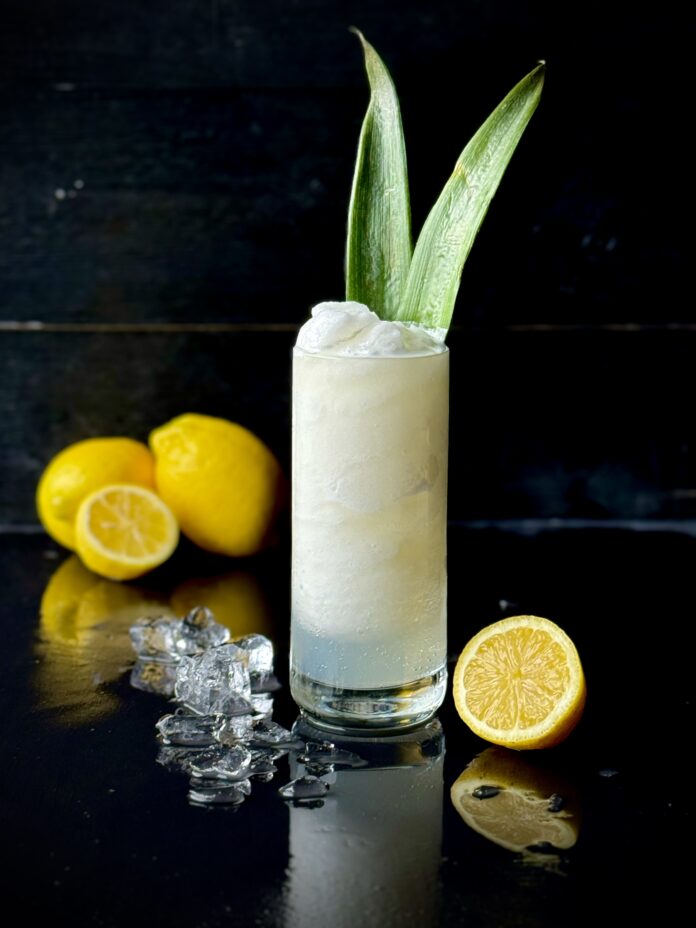 A frozen lemon cocktail in a collins glass sitting on a black reflective surface with lemons, and 2 pineapple leaves in the glass as garnish.