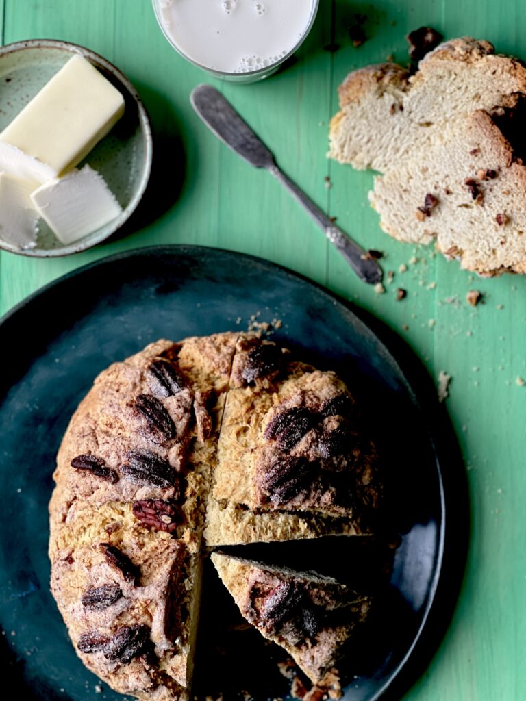 A dark green plate holds a Butter Pecan Irish Soda Bread with a slice laying on the table plus butter and a knife nearby.