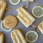 Light yellow popsicles on a textured surface covered in a Girl Scout Cookie Lemon UPS crunch coat with sliced lemons an Lemon-UPS in between the popsicles.