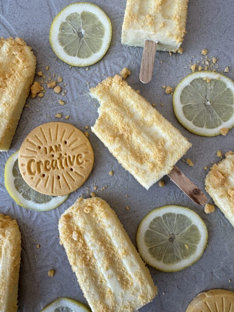 Light yellow popsicles on a textured surface covered in a Girl Scout Cookie Lemon UPS crunch coat with sliced lemons an Lemon-UPS in between the popsicles.