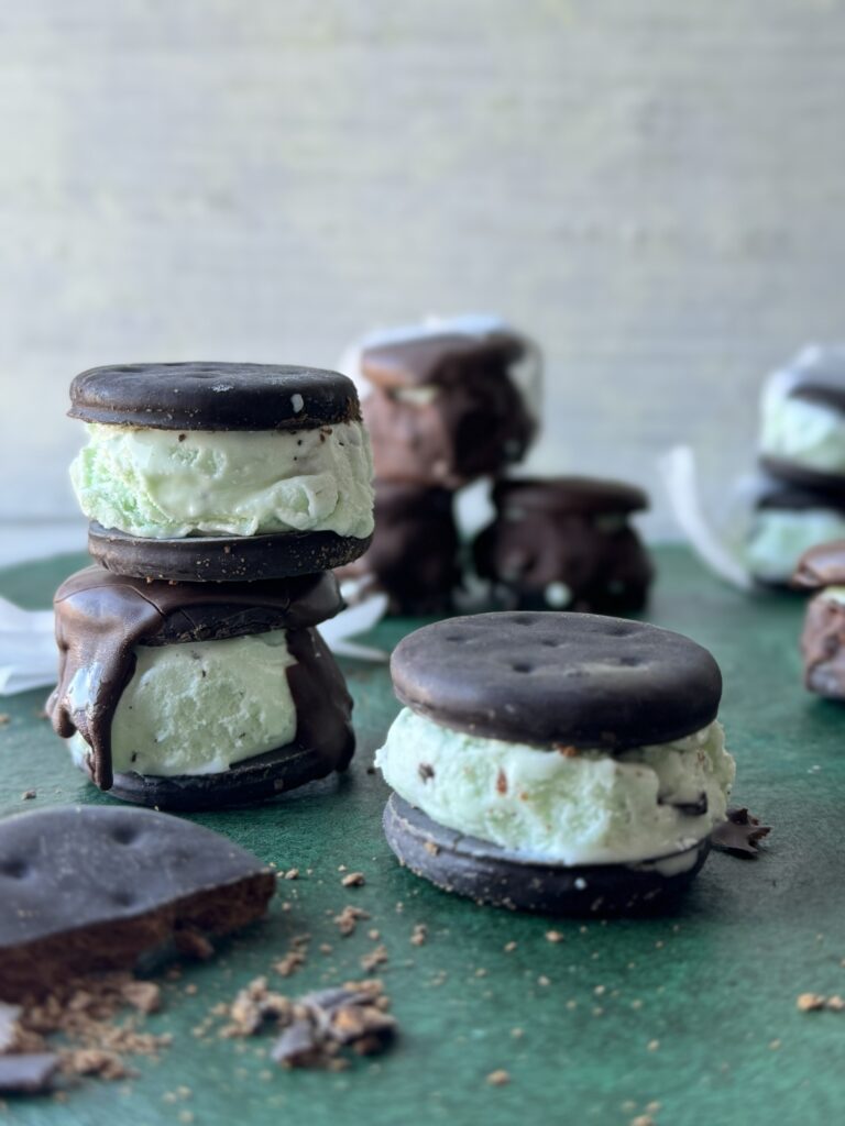 Three Thin Mint Ice Cream Sandwiches made with Thin Mint Girl Scout Cookies sit on a green table with more in the background and a few thin mint cookies scattered throughout.